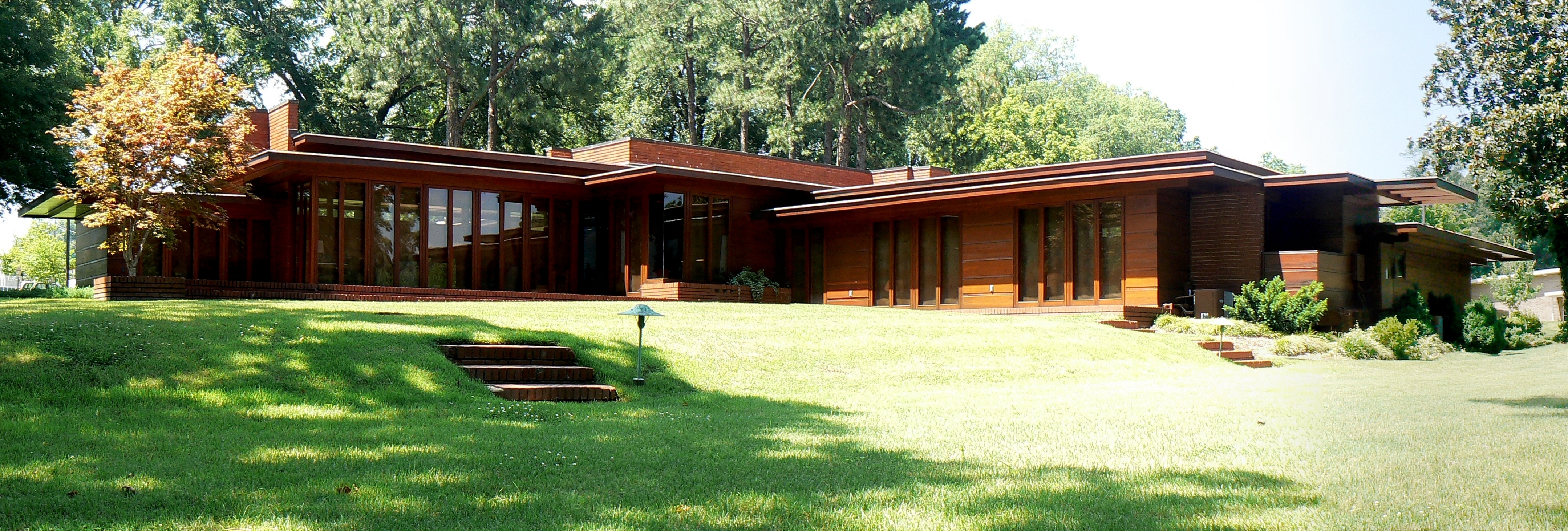 The front exterior of a usonian ranch house with floor to ceiling windows surrounded by trees and grass.